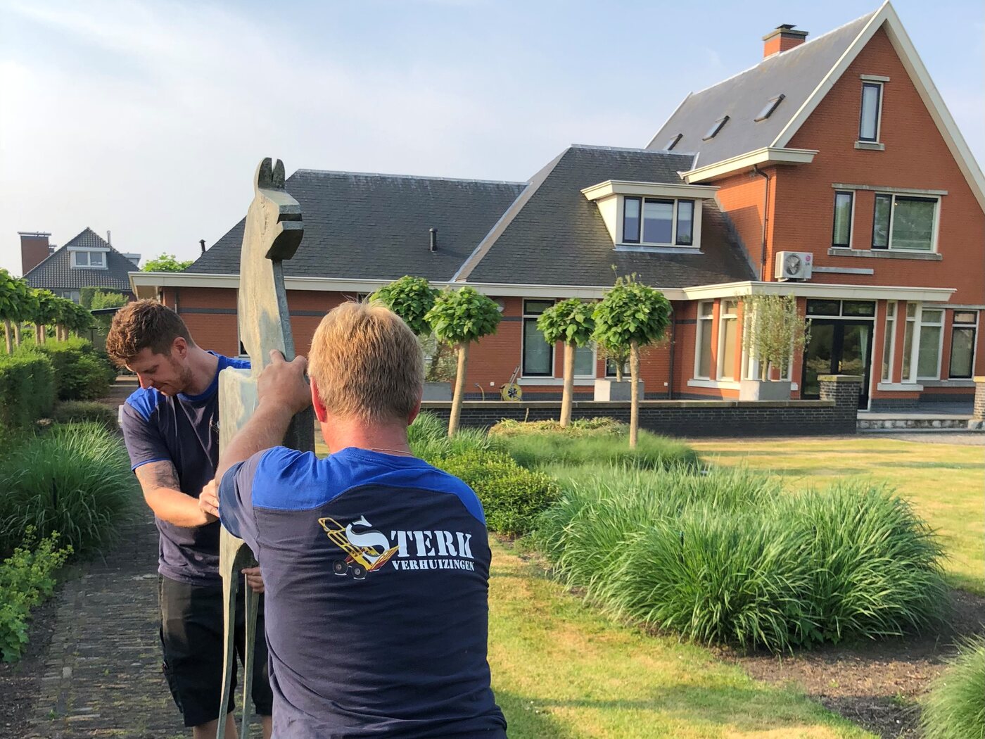 Twee verhuizers van Verhuisbedrijf Sterk Transport dragen een beeld door een tuin bij een woonhuis